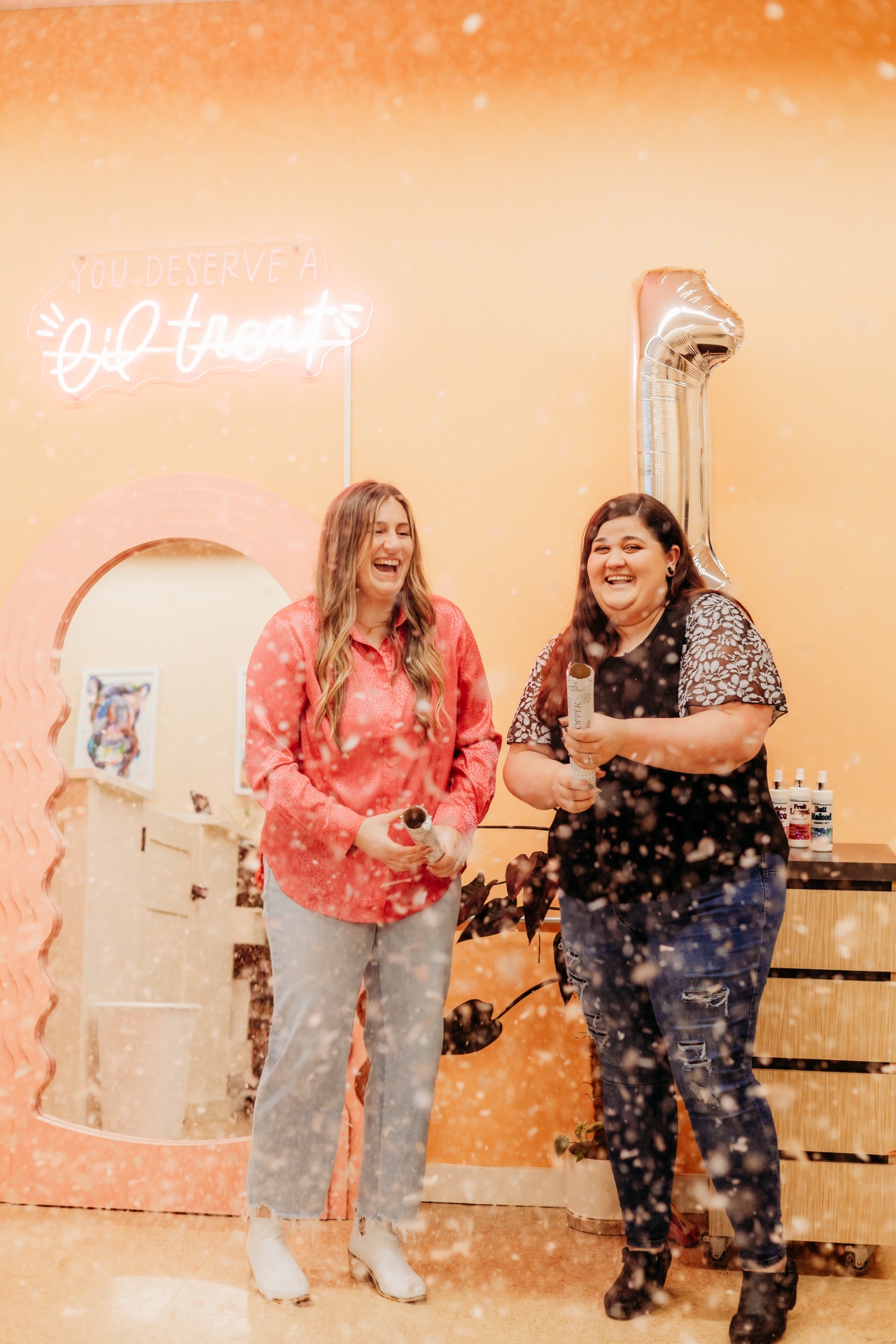Liz and Sofia laughing with confetti poppers under the salon's neon sign